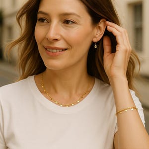 K&ouml;nnte beinhalten: Ein goldfarbenes Schmuckset bestehend aus einer Halskette, einem Armband und einem einzelnen Creolen-Ohrring, die jeweils mit kleinen, flachen, runden Pl&auml;ttchenanh&auml;ngern versehen sind, welche gleichm&auml;&szlig;ig an einer feinen Kette angeordnet sind.