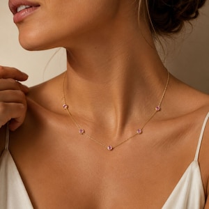 Könnte beinhalten: Eine zarte Goldkettenhalskette mit fünf gleichmäßig verteilten, runden, in Zargenfassung gefassten rosa Edelsteinen. Die Halskette liegt auf dem Schlüsselbein und zeigt eine dünne, polierte Goldgliederkette, die jede einzelne Steinfassung verbindet.