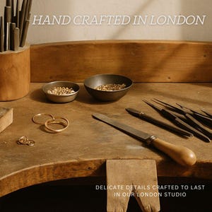 K&ouml;nnte beinhalten: Eine h&ouml;lzerne Juwelierwerkbank mit zwei kleinen Metallschalen voller Goldteile, drei Goldringen und verschiedenen Metallfeilen mit Holzgriffen. Der Text lautet HAND CRAFTED IN LONDON und DELICATE DETAILS CRAFTED TO LAST IN OUR LONDON STUDIO.