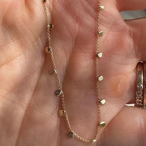 May include: A delicate gold chain necklace featuring small, evenly spaced, flat circular gold charms dangling along the length of the chain. The necklace is constructed with a fine link design, showcasing a polished metallic finish throughout.