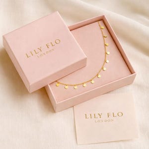 K&ouml;nnte beinhalten: Eine goldene Halskette mit kleinen runden Anh&auml;ngern in einer hellrosa quadratischen Schmuckschatulle. Der Deckel liegt neben dem Unterteil, beide mit dem goldgepr&auml;gten Text LILY FLO LONDON versehen. Eine passende Markenkarte liegt dem Set bei.
