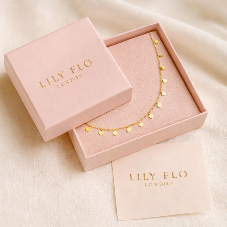 K&ouml;nnte beinhalten: Eine goldene Halskette mit kleinen runden Anh&auml;ngern in einer hellrosa quadratischen Schmuckschatulle. Der Deckel liegt neben dem Unterteil, beide mit dem goldgepr&auml;gten Text LILY FLO LONDON versehen. Eine passende Markenkarte liegt dem Set bei.