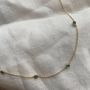 K&ouml;nnte beinhalten: Eine zarte Goldkettenhalskette mit gleichm&auml;&szlig;ig verteilten, kleinen, runden, in Zargenfassung gefassten gr&uuml;nen Edelsteinen. Die feinen Metallglieder verbinden jede kreisf&ouml;rmige Steinfassung und bilden ein schlichtes, durchgehendes Design.