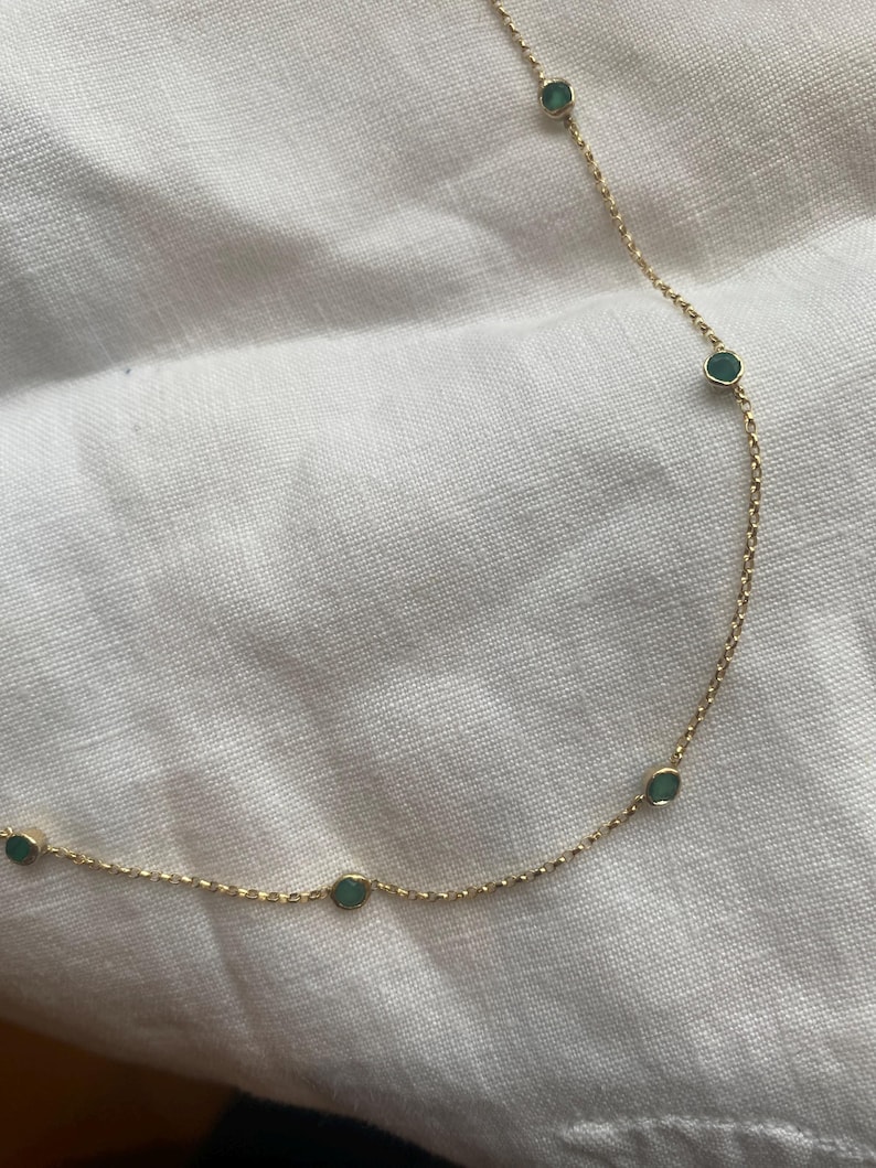 K&ouml;nnte beinhalten: Eine zarte Goldkettenhalskette mit gleichm&auml;&szlig;ig verteilten, kleinen, runden, in Zargenfassung gefassten gr&uuml;nen Edelsteinen. Die feinen Metallglieder verbinden jede kreisf&ouml;rmige Steinfassung und bilden ein schlichtes, durchgehendes Design.