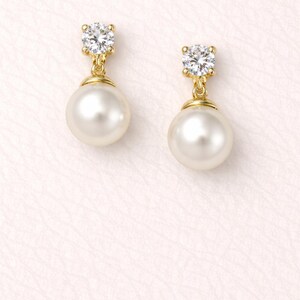 May include: A pair of gold drop earrings featuring a round white pearl suspended from a single clear, faceted round-cut stone set in a gold prong setting. The design combines the luster of the pearl with the sparkle of the clear stone.