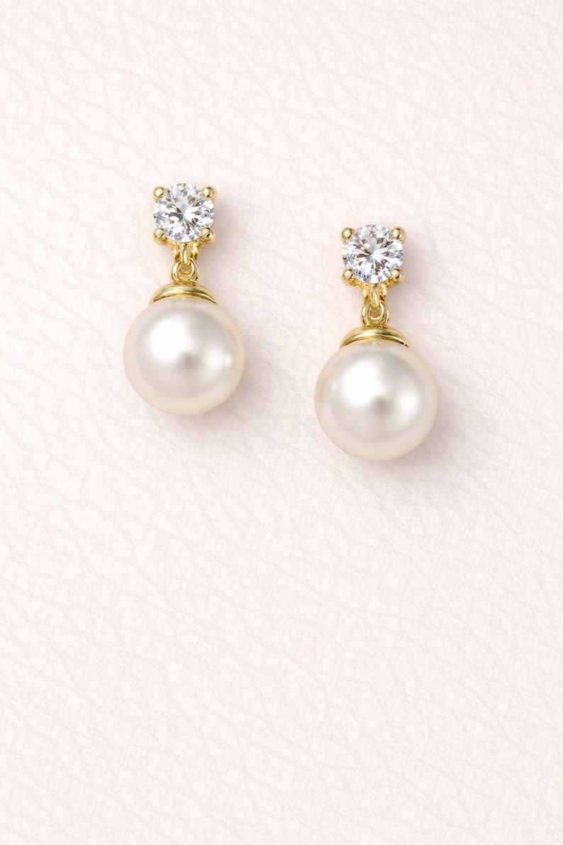 May include: A pair of gold drop earrings featuring a round white pearl suspended from a single clear, faceted round-cut stone set in a gold prong setting. The design combines the luster of the pearl with the sparkle of the clear stone.