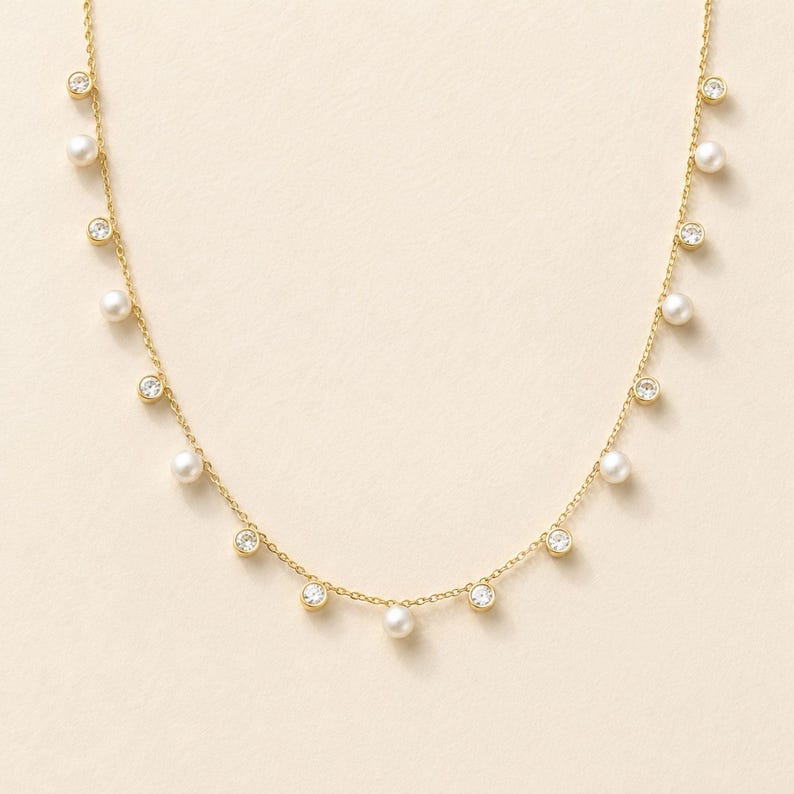May include: A gold chain necklace featuring a delicate arrangement of alternating round white pearls and clear, bezel-set circular crystals suspended along the length of the chain, creating a balanced and symmetrical jewelry design.