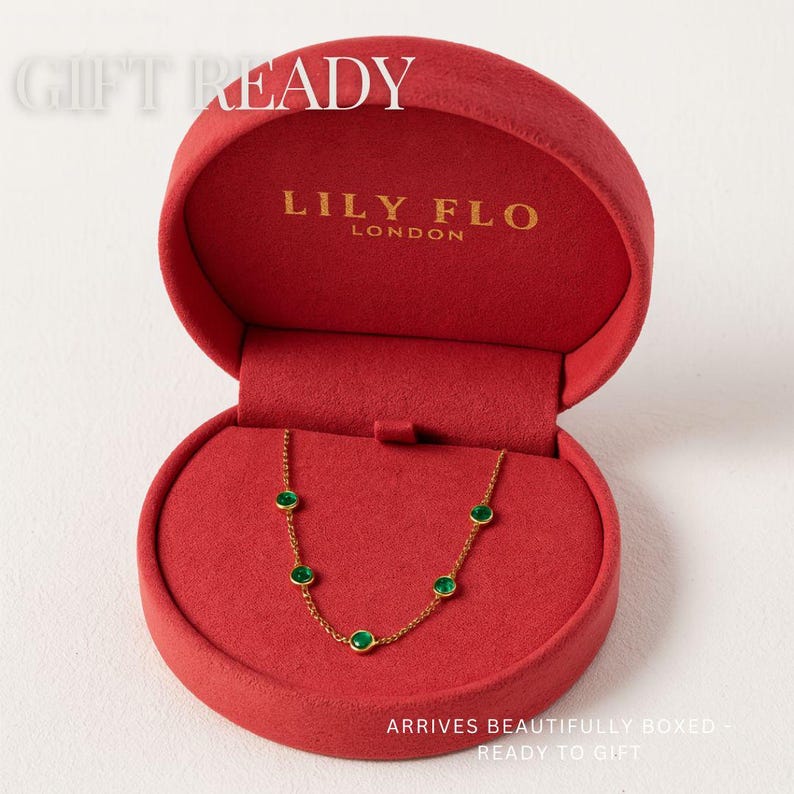 May include: A gold chain necklace featuring five small, round, green bezel-set stones, presented in a round red velvet jewelry box. The interior lid is printed with the text LILY FLO LONDON and the text GIFT READY is visible at the top.