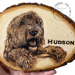 Op de afbeelding: Een houten schijf met een ingebrand beeld van een bruine hond met de naam "HUDSON" gebrand onder het beeld. De houten schijf heeft een natuurlijke schorsrand en is lichtbruin.