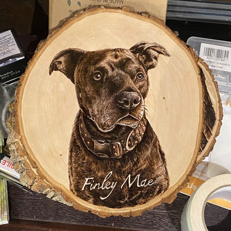 Op de afbeelding: Een houten schijf met een gebrand beeld van een zwarte hond met een bruine halsband. De naam van de hond, Finley Mae, staat onder de afbeelding.