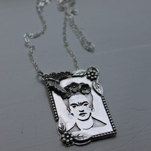 Puede incluir: Un collar de plata con un colgante rectangular que presenta un retrato de Frida Kahlo con una mariposa y detalles florales.