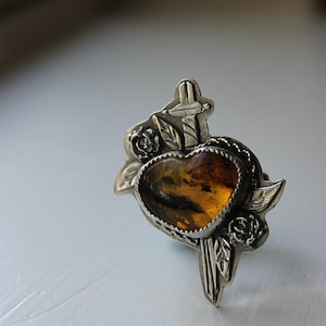 Puede incluir: Anillo plateado con una piedra de ámbar en forma de corazón. El anillo tiene un diseño decorativo con rosas, hojas y una cruz. La piedra de ámbar es de color marrón dorado.