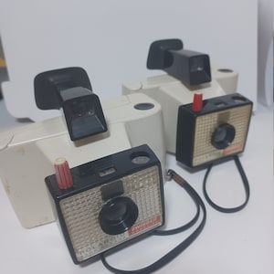 May include: Two vintage white Polaroid Swinger Land Cameras with black and red accents. The cameras have a black strap and the text "POLAROID LAND CAMERA" and "SWINGER" is printed on the front.