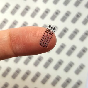 Op de afbeelding: Een transparante sticker met een zwarte omtrek van een afstandsbediening. De sticker zit op een vinger.