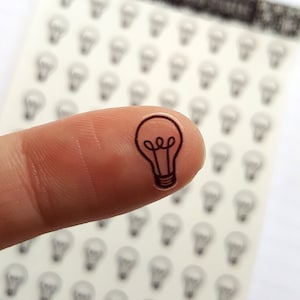 May include: A sheet of clear stickers with a black outline of a light bulb. A finger is holding one of the stickers.