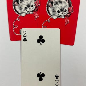 May include: A playing card with the number two of clubs, with two red cards featuring a kitten illustration. The kitten has gray and white fur, a red bow, and is hanging from a string. The cards are on a white background.