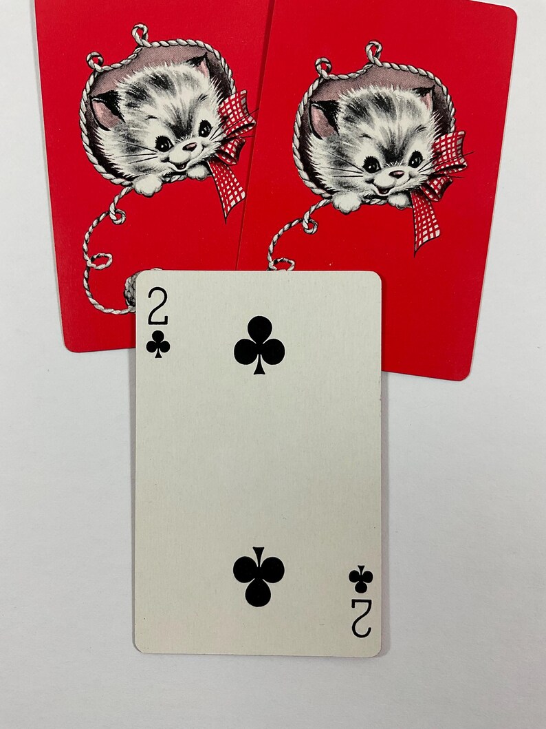 May include: A playing card with the number two of clubs, with two red cards featuring a kitten illustration. The kitten has gray and white fur, a red bow, and is hanging from a string. The cards are on a white background.