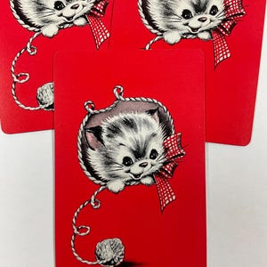 May include: Red playing cards featuring a vintage illustration of a gray and white kitten with a red and white checkered bow. The kitten is inside a rope border and is connected to a ball of yarn.