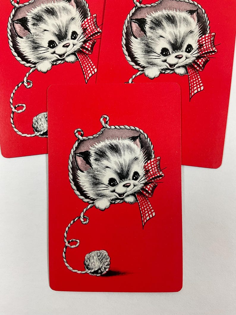 May include: Red playing cards featuring a vintage illustration of a gray and white kitten with a red and white checkered bow. The kitten is inside a rope border and is connected to a ball of yarn.