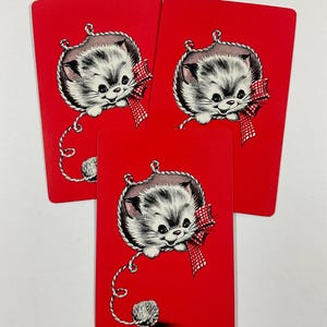 May include: Three red playing cards featuring a vintage illustration of a gray and white kitten with a red bow and a ball of yarn. The kitten is hanging from a rope. The cards are arranged in a fan shape.