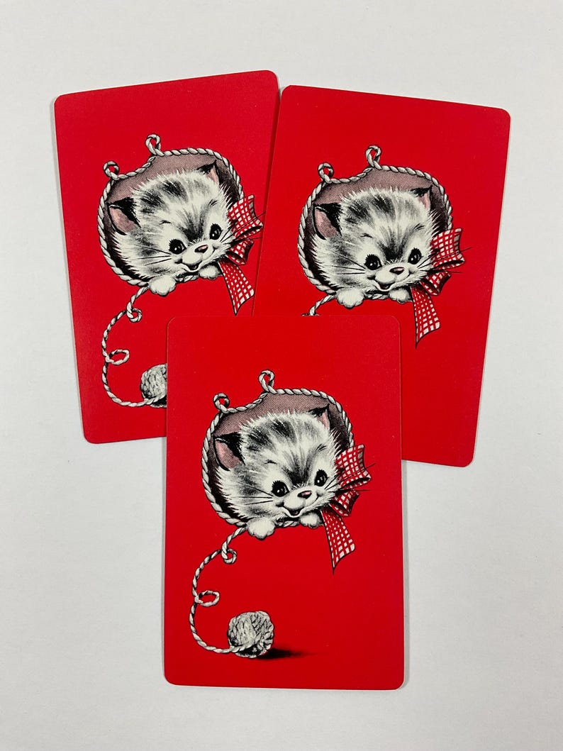 May include: Three red playing cards featuring a vintage illustration of a gray and white kitten with a red bow and a ball of yarn. The kitten is hanging from a rope. The cards are arranged in a fan shape.