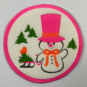 May include: A circular Christmas decoration featuring a snowman with a pink top hat and orange bow tie. The snowman is surrounded by green Christmas trees and a pink border. The snowman is pulling a sled with a Christmas tree.