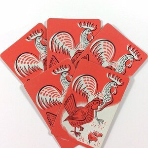 May include: Five red playing cards with white and red rooster illustrations. The cards are stacked in a fan shape, with the top card showing a rooster with a red comb and wattles, and two smaller roosters below.