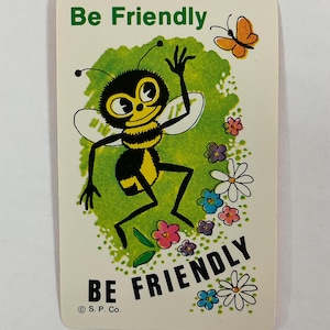 Op de afbeelding: Een vintage kaart met een cartoon bijenillustratie. De bij is zwart en geel, zwaaiend met een vriendelijke uitdrukking. De kaart heeft de woorden "Be Friendly" in groen en zwart, met een vlinder en bloemen.