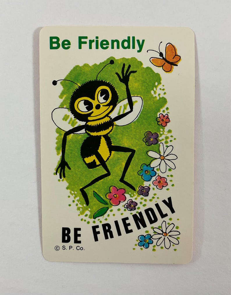Op de afbeelding: Een vintage kaart met een cartoon bijenillustratie. De bij is zwart en geel, zwaaiend met een vriendelijke uitdrukking. De kaart heeft de woorden "Be Friendly" in groen en zwart, met een vlinder en bloemen.