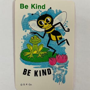Op de afbeelding: Een vintage kaart met een cartoon illustratie van een bij en een kikker. De bij is zwart en geel, en de kikker is groen. De kaart heeft de woorden "Be Kind" in groene en zwarte tekst.
