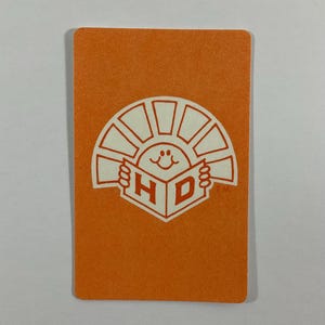 Op de afbeelding: Oranje rechthoekige kaart met een wit logo met een lachende zon die een boek vasthoudt met de letters "H" en "D". De kaart heeft afgeronde hoeken en een getextureerd oppervlak.