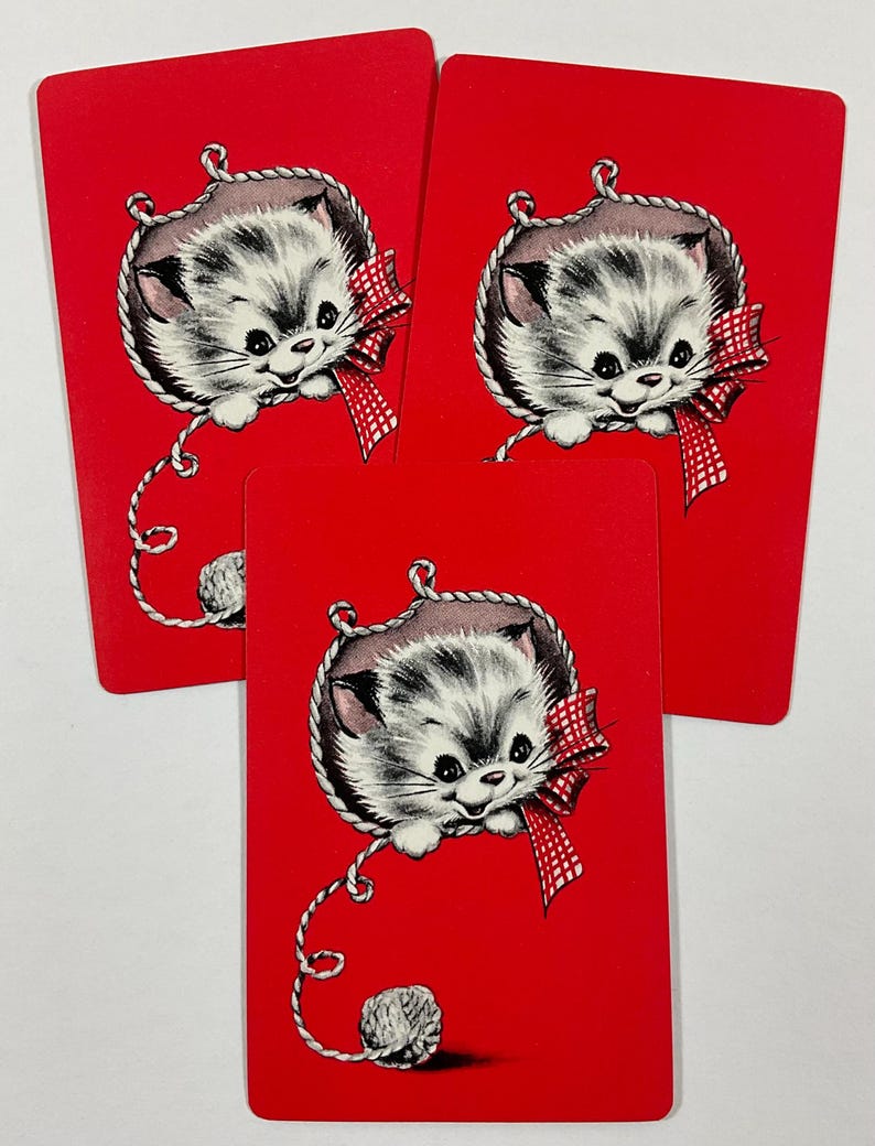 May include: Three red playing cards featuring a cute kitten illustration. The kitten is gray and white, wearing a red and white checkered bow, and surrounded by a rope border. A ball of yarn is at the bottom.