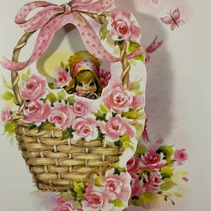 May include: A vintage-style greeting card featuring a woven basket overflowing with pink roses. A young girl peeks out from behind the flowers. Pink butterflies and a pink ribbon bow add to the floral theme. The card has a 3D effect.