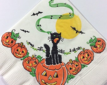 May include: White paper napkin with a Halloween theme. It features a black cat sitting on a jack-o-lantern, surrounded by other jack-o-lanterns. There is a yellow moon in the background with green musical notes and bats flying around.