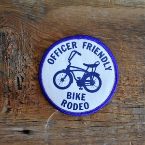 Może przedstawiać: Okrągła naszywka w kolorze białym i niebieskim z fioletowym obramowaniem. Naszywka przedstawia sylwetkę roweru z napisem "Officer Friendly Bike Rodeo".