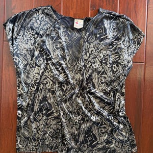 May include: A short-sleeved top featuring a black base with a metallic silver abstract pattern. The garment has a wide, rounded neckline and a loose, draped fit. A small white rectangular tag is visible at the inner neckline with black text.