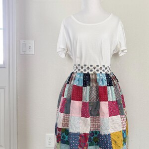 May include: A white top and a patchwork skirt. The skirt is made of squares of fabric in various colours, including red, blue, pink, and yellow. The skirt is displayed on a mannequin.