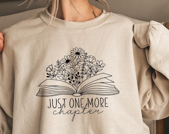 Peut inclure: Sweat-shirt beige avec un motif noir d'un livre ouvert orné de fleurs et le texte "JUST ONE MORE chapter". Le sweat-shirt est de couleur neutre et a une coupe décontractée.