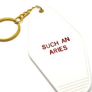 May include: A white retro-style keychain with the text "SUCH AN ARIES" in red. The keychain has a gold-colored chain and key ring. The design is simple and the keychain is likely made of plastic.