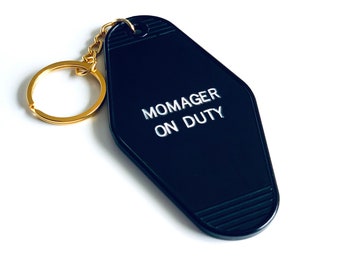 Può includere: Portachiavi nero a forma di chiave di motel con un anello di chiave dorato. Il portachiavi ha il testo "Momager on Duty" in bianco.