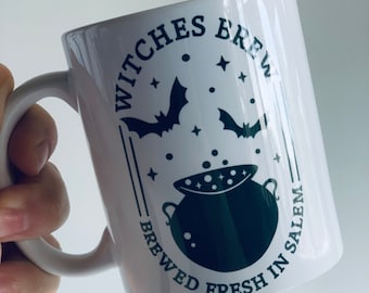 May include: White ceramic mug with a green design. The text reads "Witches Brew, Brewed Fresh in Salem." The design includes bats, stars, and a bubbling cauldron. The mug has a curved handle.