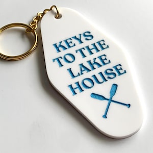 May include: White keychain with gold-colored hardware. The keychain has the text "KEYS TO THE LAKE HOUSE" in blue, along with a blue crossed oars graphic.