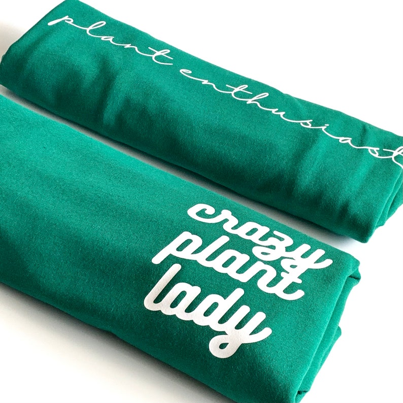 May include: Two green t-shirts with white text. The top shirt says "plant enthusiast" and the bottom shirt says "crazy plant lady".