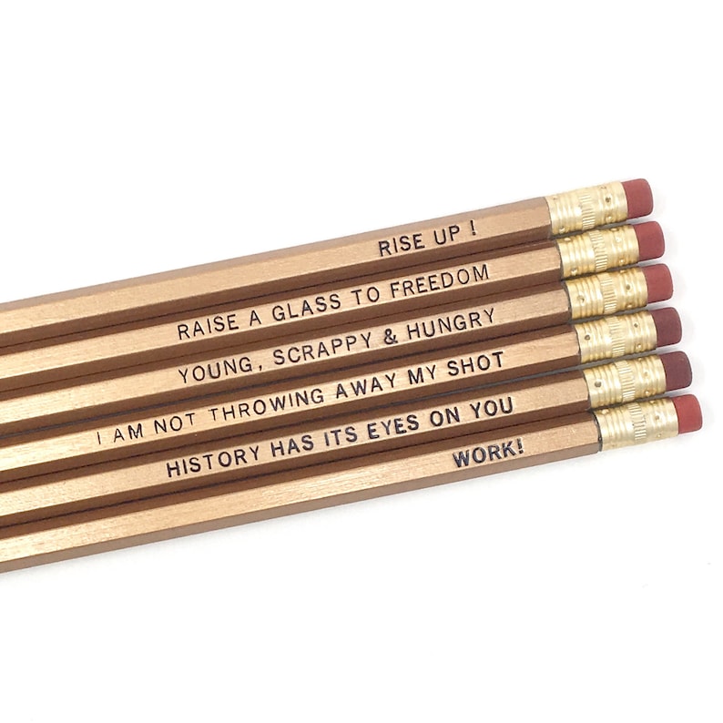 May include: A set of six gold pencils with inspirational quotes engraved on them. The quotes are: "Rise Up!", "Raise a Glass to Freedom", "Young, Scrappy & Hungry", "I am not throwing away my shot", "History has its eyes on you", and "Work!"
