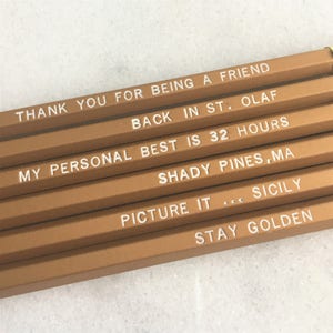 May include: Set of six gold pencils with white text. The text on the pencils reads: "Thank you for being a friend", "Back in St. Olaf", "My personal best is 32 hours", "Shady Pines, MA", "Picture it... Sicily", and "Stay Golden".