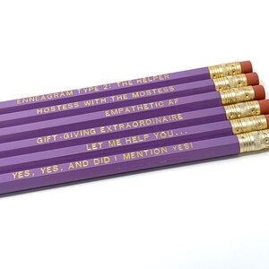 Può includere: Sei matite viola con scritte dorate. Le matite presentano frasi come "Enneagramma Tipo 2: L'aiutante" e "Hostess with the Mostess". Ogni matita ha una gomma rosa e un puntale in metallo dorato.