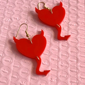 May include: A pair of red heart-shaped earrings with gold hooks and devil horns. The earrings have a tail that ends in an arrow.