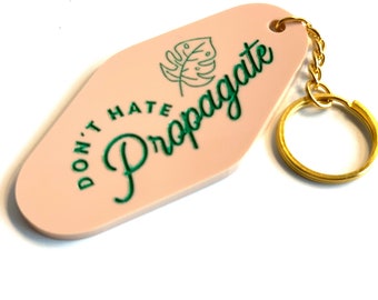 Pode incluir: Um chaveiro rosa estilo motel com as palavras "DON'T HATE Propagate" em verde. Um desenho de folha verde está acima da palavra "Propagate". O chaveiro tem uma corrente e um porta-chaves dourados.