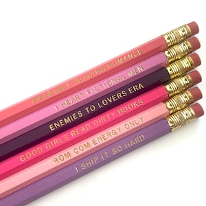 May include: A collection of seven pencils in shades of pink and purple, each with gold lettering. The pencils have phrases like "Probably Reading Romance" and "I Heart Fictional Men". Each pencil has a pink eraser and gold ferrule.