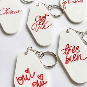 Puede incluir: Cinco llaveros blancos con letras rojas. Los llaveros tienen forma de llaves de motel y presentan las frases "Cherie", "C'est la Vie", "Je t'aime", "Tres Bien" y "Qui ou".